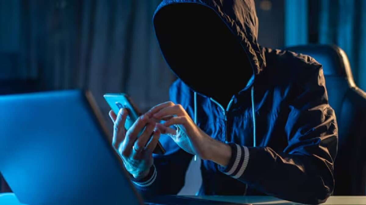 The image of a person in a dark hoodie with a hidden face sits at a desk, using a smartphone and a laptop in a dimly lit room with blue lighting, suggesting a secretive or cyber activity.