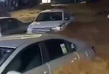 A nighttime scene in Asir Province, Saudi Arabia, showing several cars partially submerged in muddy floodwater on a street, with a person in traditional white clothing standing nearby.