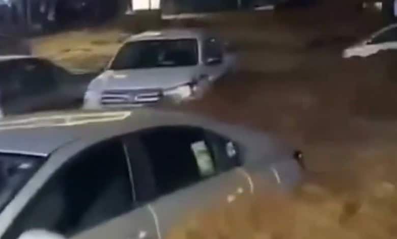 A nighttime scene in Asir Province, Saudi Arabia, showing several cars partially submerged in muddy floodwater on a street, with a person in traditional white clothing standing nearby.