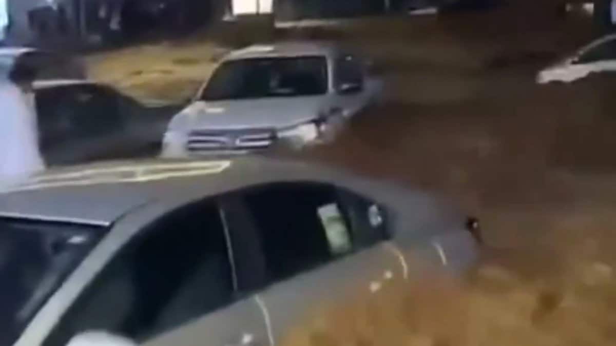 A nighttime scene in Asir Province, Saudi Arabia, showing several cars partially submerged in muddy floodwater on a street, with a person in traditional white clothing standing nearby.