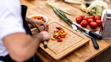 Chef chopping fresh cherry tomatoes on a wooden board with vegetables and kitchen tools around.
