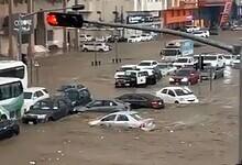 Representative image of a flooded street in Makkah, with cars and a bus partially submerged in muddy water, surrounded by buildings and traffic lights showing red.