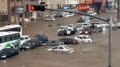 Representative image of a flooded street in Makkah, with cars and a bus partially submerged in muddy water, surrounded by buildings and traffic lights showing red.