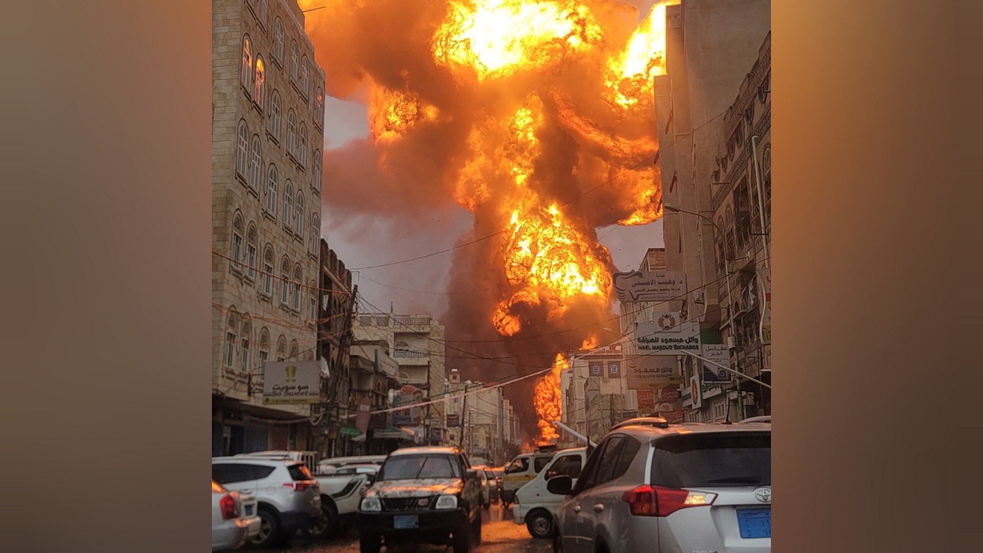 A massive explosion with flames and smoke rises in a city street lined with buildings and cars, suggesting a military strike.