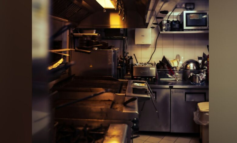 A busy kitchen with stainless steel appliances, a microwave, pots, pans, and a sink, illuminated by warm lighting.