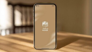 The image of a smartphone on a wooden table showing the Nusuk app splash screen with Arabic logo on a beige background.