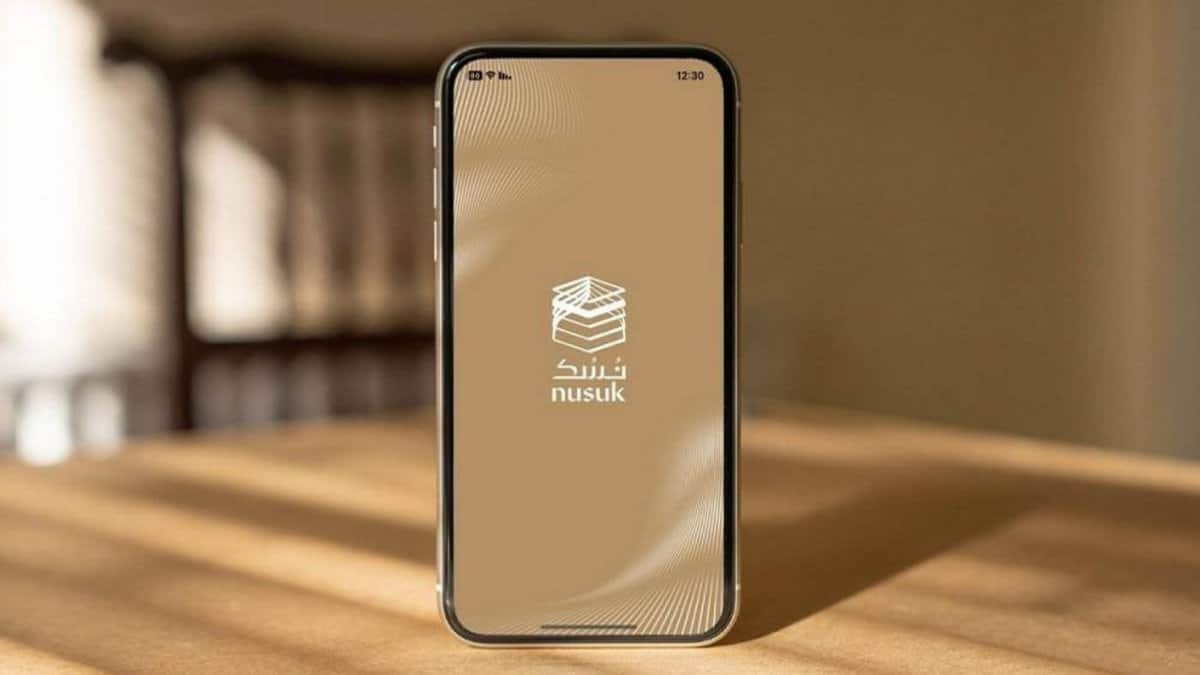 The image of a smartphone on a wooden table showing the Nusuk app splash screen with Arabic logo on a beige background.