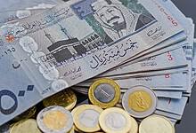 A stack of Saudi Riyal banknotes and coins, featuring denominations like 500 and 1 riyal, with the Kaaba image and Arabic script on the notes, and gold/silver coins, on a dark surface.