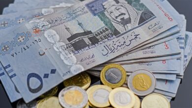A stack of Saudi Riyal banknotes and coins, featuring denominations like 500 and 1 riyal, with the Kaaba image and Arabic script on the notes, and gold/silver coins, on a dark surface.