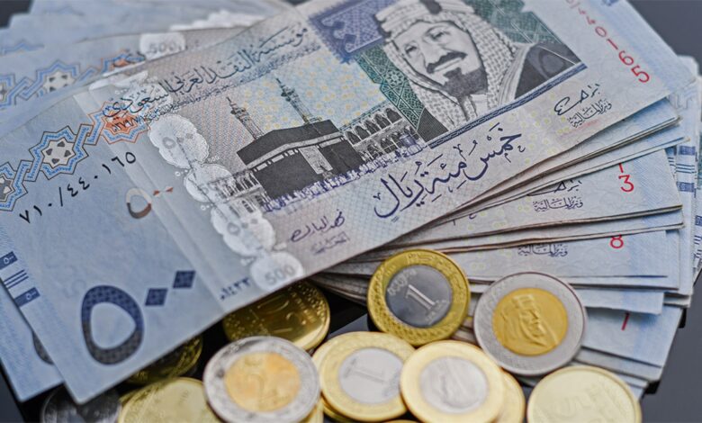 A stack of Saudi Riyal banknotes and coins, featuring denominations like 500 and 1 riyal, with the Kaaba image and Arabic script on the notes, and gold/silver coins, on a dark surface.