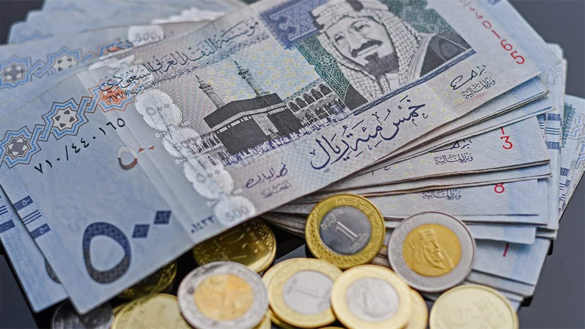 A stack of Saudi Riyal banknotes and coins, featuring denominations like 500 and 1 riyal, with the Kaaba image and Arabic script on the notes, and gold/silver coins, on a dark surface.