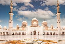 A stunning view of the Sheikh Zayed Grand Mosque in Abu Dhabi, featuring its grand white marble architecture, multiple domes, and tall minarets under a partly cloudy blue sky. The intricate floral mosaic on the courtyard floor adds vibrant color to the scene