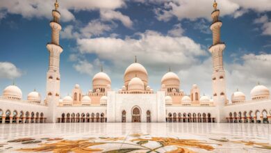 A stunning view of the Sheikh Zayed Grand Mosque in Abu Dhabi, featuring its grand white marble architecture, multiple domes, and tall minarets under a partly cloudy blue sky. The intricate floral mosaic on the courtyard floor adds vibrant color to the scene