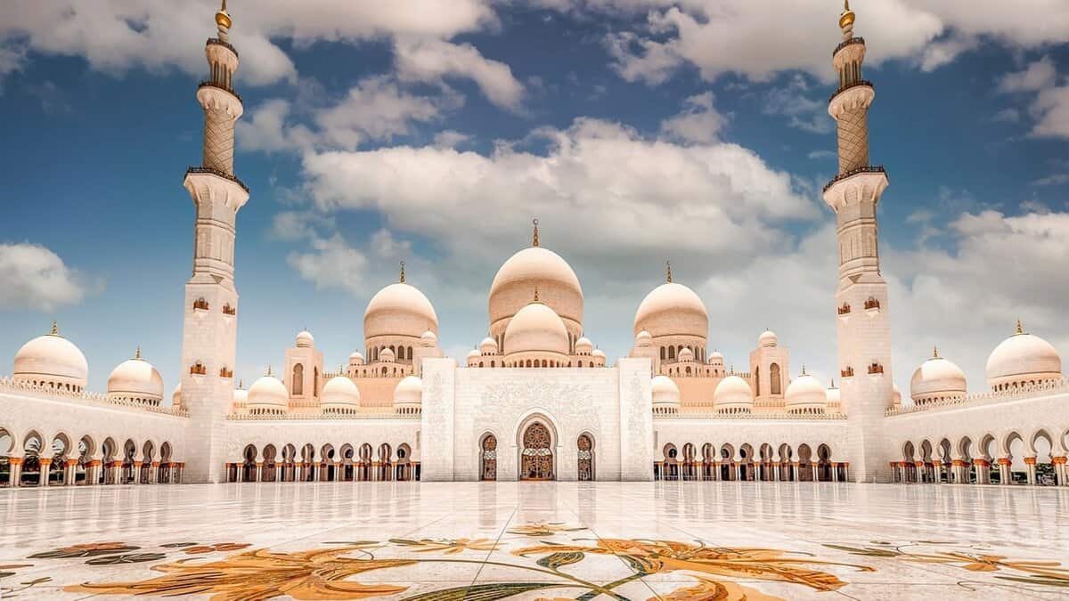 A stunning view of the Sheikh Zayed Grand Mosque in Abu Dhabi, featuring its grand white marble architecture, multiple domes, and tall minarets under a partly cloudy blue sky. The intricate floral mosaic on the courtyard floor adds vibrant color to the scene