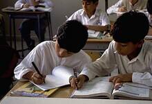 Two students in white uniforms writing in notebooks at desks in a UAE school classroom, with other students working in the background.