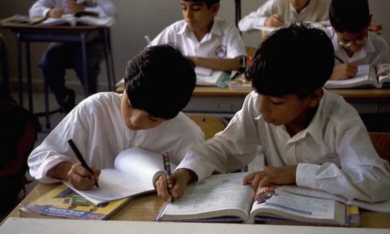 Two students in white uniforms writing in notebooks at desks in a UAE school classroom, with other students working in the background.