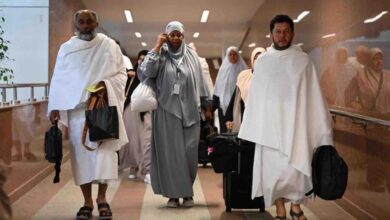 A group of Umrah pilgrims, dressed in Ihram and modest attire, walking through an airport corridor in Saudi Arabia. Some carry luggage and bags, with a modern interior featuring glass railings and overhead lighting.