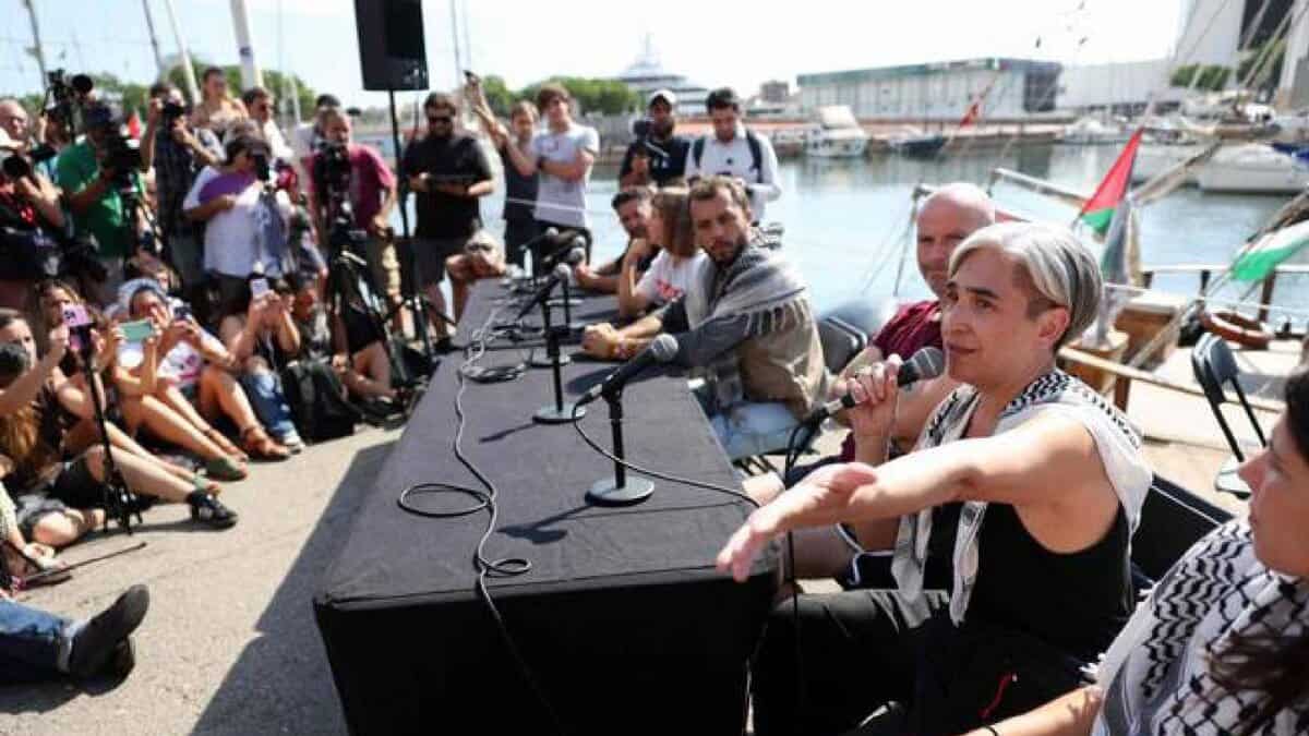 Former Barcelona mayor Ada Colau speaks at a press conference during the launch of a civilian flotilla in Barcelona, with media and supporters gathered around.