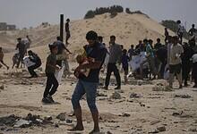 A crowded scene of people, including a man holding a child, walking across a debris-strewn sandy area, seeking aid in a conflict zone.