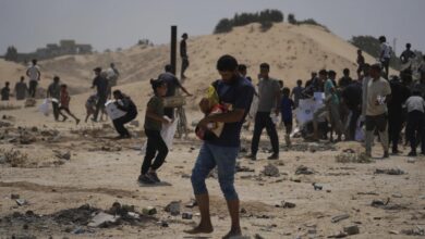 A crowded scene of people, including a man holding a child, walking across a debris-strewn sandy area, seeking aid in a conflict zone.