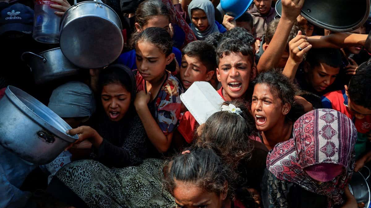 A crowded scene of people, mostly children, holding pots and containers, reaching out in a desperate manner.