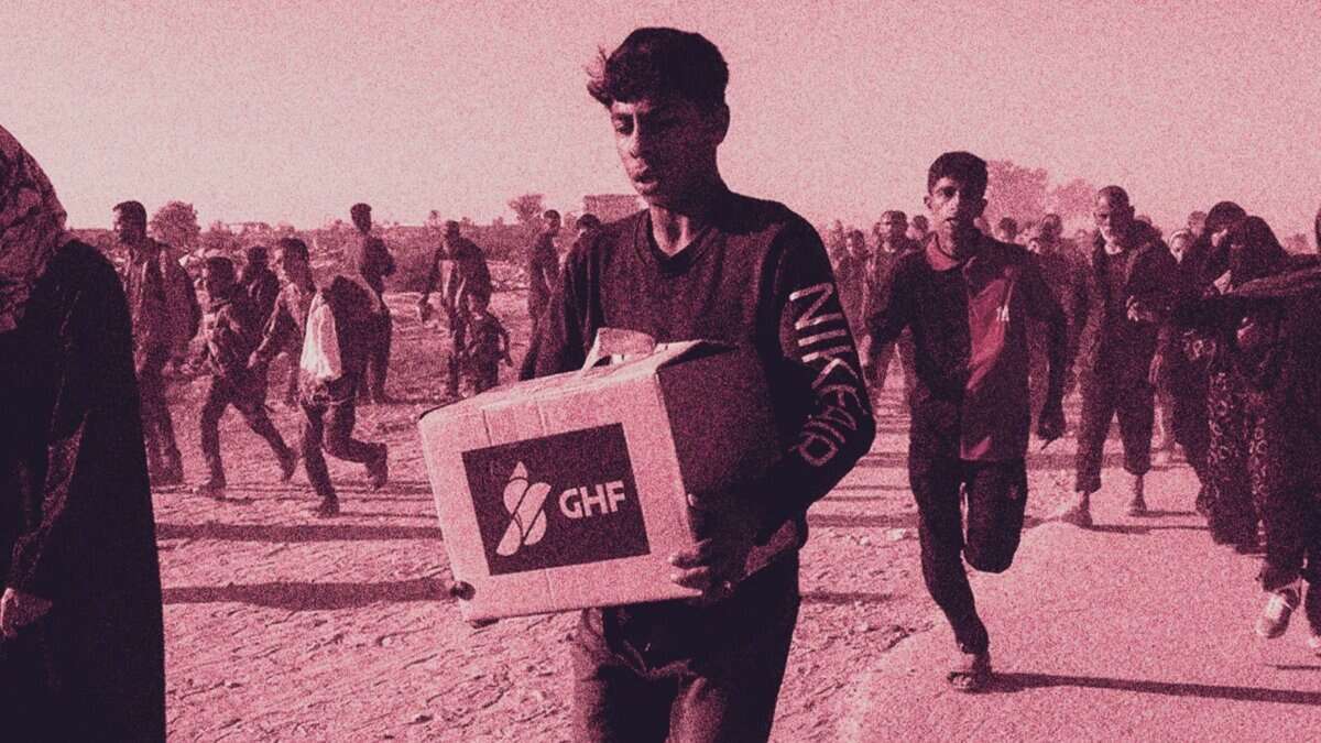 A young Palestinian man carries a Gaza Humanitarian Foundation (GHF) aid box while others walk and run behind him in a crowded, dusty area.