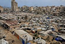 A sprawling refugee camp in Gaza with numerous tents and makeshift shelters, surrounded by damaged buildings and a tall, partially destroyed high-rise, under a cloudy sky.