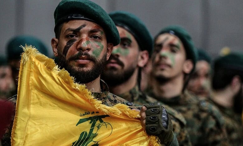 A Hezbollah militant holds the group's yellow flag during a parade, with fellow fighters in camouflage and green berets standing behind him.