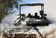 A military tank moving through a dusty environment with two soldiers visible in the turret.