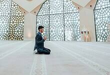 Man praying inside a mosque, sitting on carpet with hands raised in supplication.