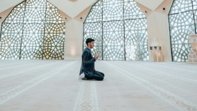 Man praying inside a mosque, sitting on carpet with hands raised in supplication.