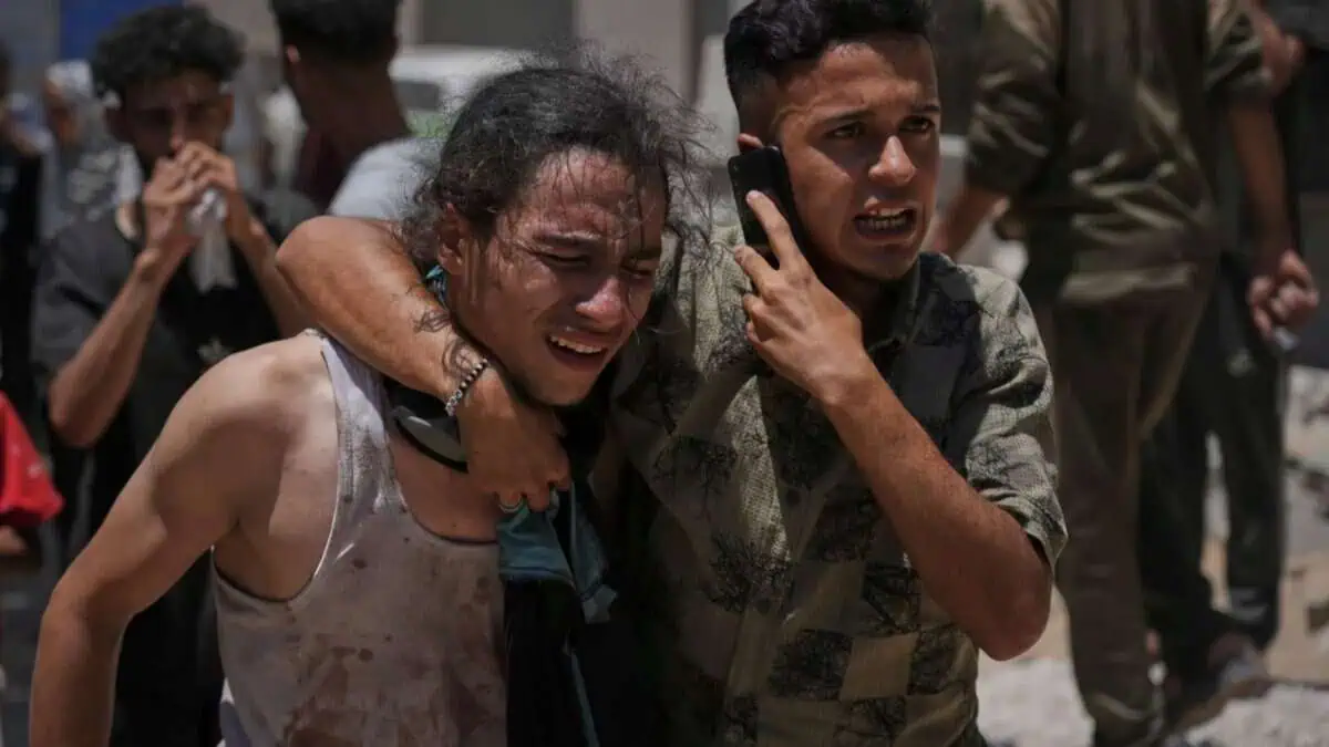 Two distressed Palestinians, one supporting the other, react emotionally while carrying a body at Shifa Hospital in Gaza City after an aid truck incident.