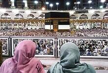 Two people in colorful headscarves observe a large crowd gathered around the Kaaba at night in a grand mosque.