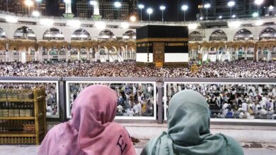 Two people in colorful headscarves observe a large crowd gathered around the Kaaba at night in a grand mosque.