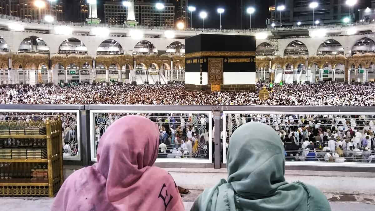 Two people in colorful headscarves observe a large crowd gathered around the Kaaba at night in a grand mosque.