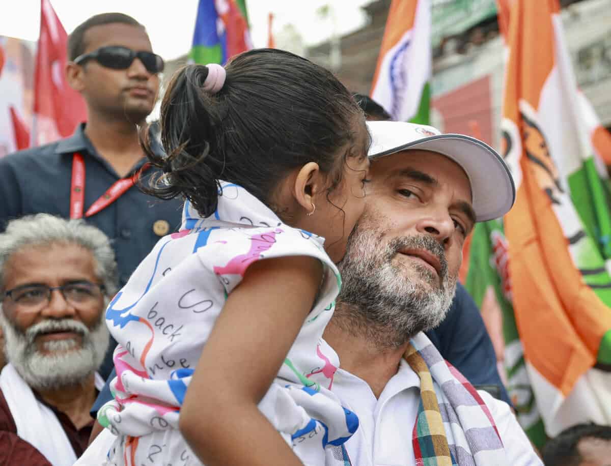 Child whispering to man at political rally, India election campaign, vibrant flags in background.