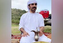 A Saudi man dressed in traditional white attire and head covering sits outdoors on a patterned carpet, holding a traditional Arabic coffee pot.