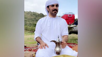 A Saudi man dressed in traditional white attire and head covering sits outdoors on a patterned carpet, holding a traditional Arabic coffee pot.