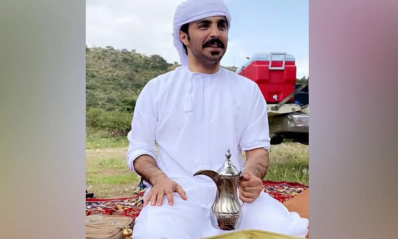 A Saudi man dressed in traditional white attire and head covering sits outdoors on a patterned carpet, holding a traditional Arabic coffee pot.