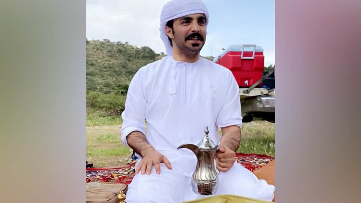 A Saudi man dressed in traditional white attire and head covering sits outdoors on a patterned carpet, holding a traditional Arabic coffee pot.