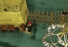 Overhead view of a person in a red cap and dark clothing near a yellow structure, with chairs and a chandelier in the background, inside a mosque.