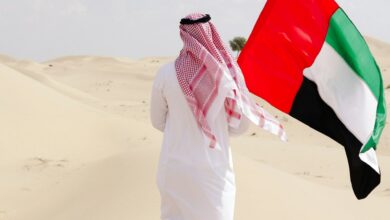 Man in traditional Emirati attire holding UAE flag in desert.