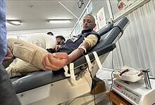 A Palestinian man with a beard lies reclined on a medical bed in a Gaza blood bank, donating blood through an IV in his arm; a nurse in white adjusts the tube connected to a blood collection bag on a scale, with Arabic health posters and heart symbols on the walls.