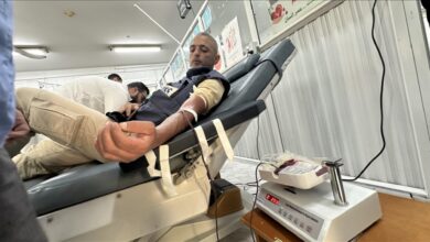 A Palestinian man with a beard lies reclined on a medical bed in a Gaza blood bank, donating blood through an IV in his arm; a nurse in white adjusts the tube connected to a blood collection bag on a scale, with Arabic health posters and heart symbols on the walls.