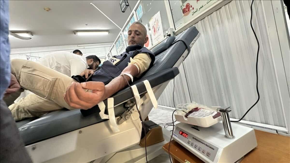 A Palestinian man with a beard lies reclined on a medical bed in a Gaza blood bank, donating blood through an IV in his arm; a nurse in white adjusts the tube connected to a blood collection bag on a scale, with Arabic health posters and heart symbols on the walls.