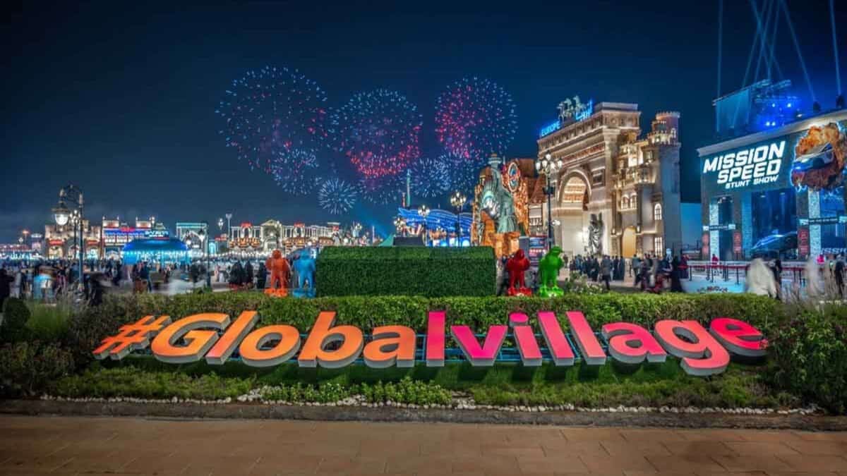 A night view of Dubai’s Global Village with fireworks lighting up the sky, crowds walking around brightly lit pavilions, and a large #GlobalVillage sign in the foreground.