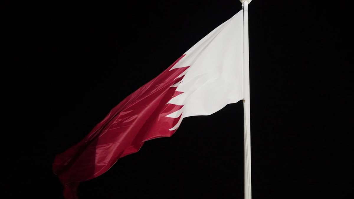 Qatar’s national flag waving against a dark night sky. The maroon-and-white flag is mounted on a tall pole.