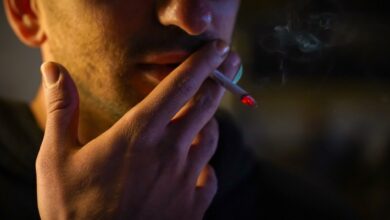 Close-up of a man smoking a cigarette, with the glowing red tip of the cigarette clearly visible. His hand is holding the cigarette near his lips, and faint smoke is rising in the background.