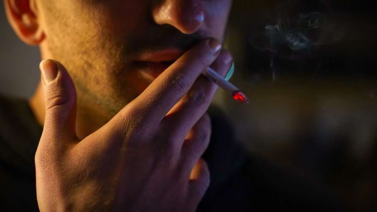 Close-up of a man smoking a cigarette, with the glowing red tip of the cigarette clearly visible. His hand is holding the cigarette near his lips, and faint smoke is rising in the background.