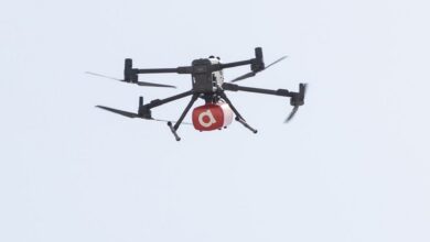 A delivery drone flying in the sky, carrying a red package with a white letter "a" on it.